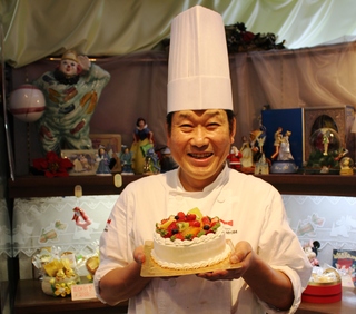 ホールケーキを手に持ち笑顔のバロンミヨシさんの写真